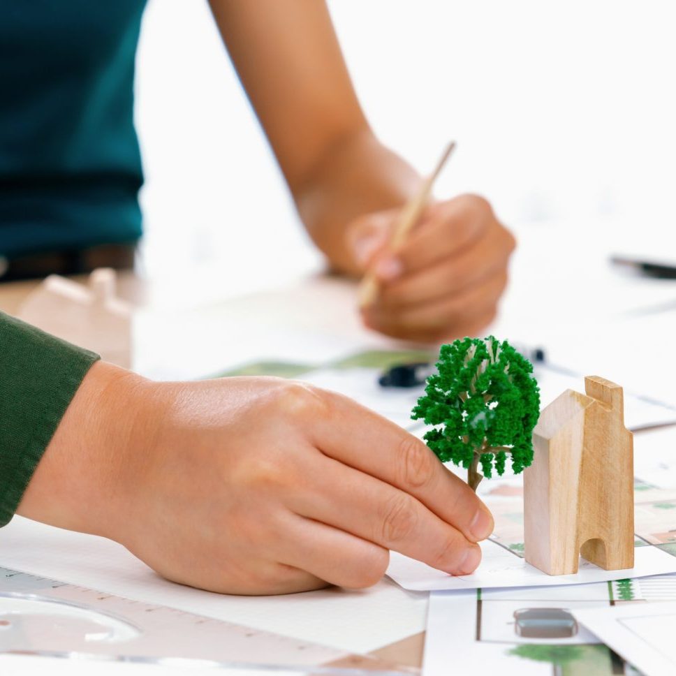 Professional architect team discuss about green design of eco house on meeting table with blueprint and model scatter around. Closeup. Focus on hand. Green city concept. Delineation.